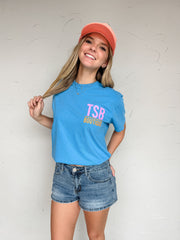 Person wearing a blue t-shirt with 'TSB Boutique' text, denim shorts, and a pink cap against a white wall.