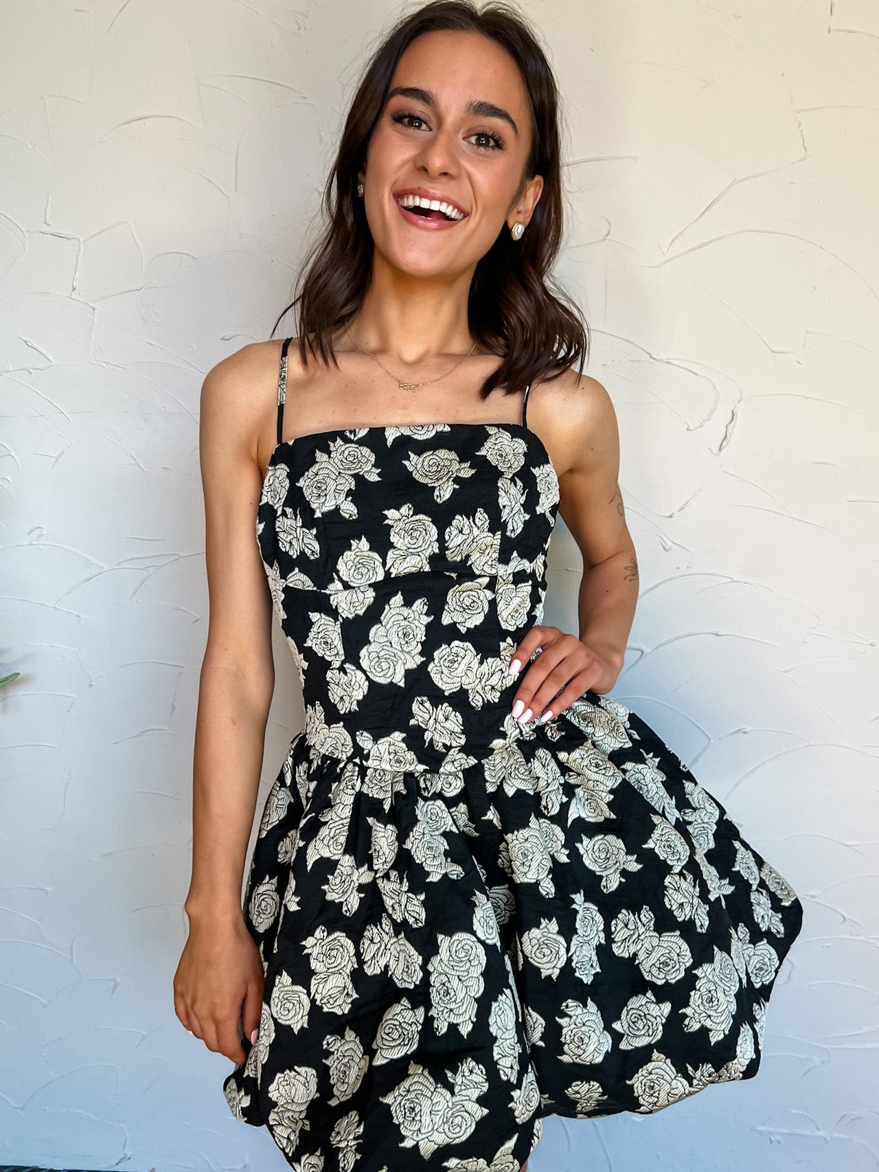 Woman wearing a black floral dress against a textured white wall.