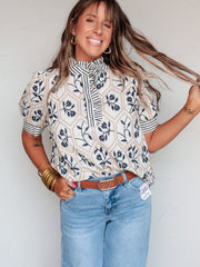 A waist-up photograph of a smiling woman with medium-length brown hair and bangs holding a strand of her hair while posing against a textured white wall. She is wearing a cream-colored short-sleeved blouse made from 67% Tencel, 21% nylon, and 12% linen fabric featuring navy floral print, a high ruffled collar, striped trim, and puff sleeves, tucked into light-wash denim jeans with a cognac brown belt and styled with gold hoop earrings and stacked gold bangles.