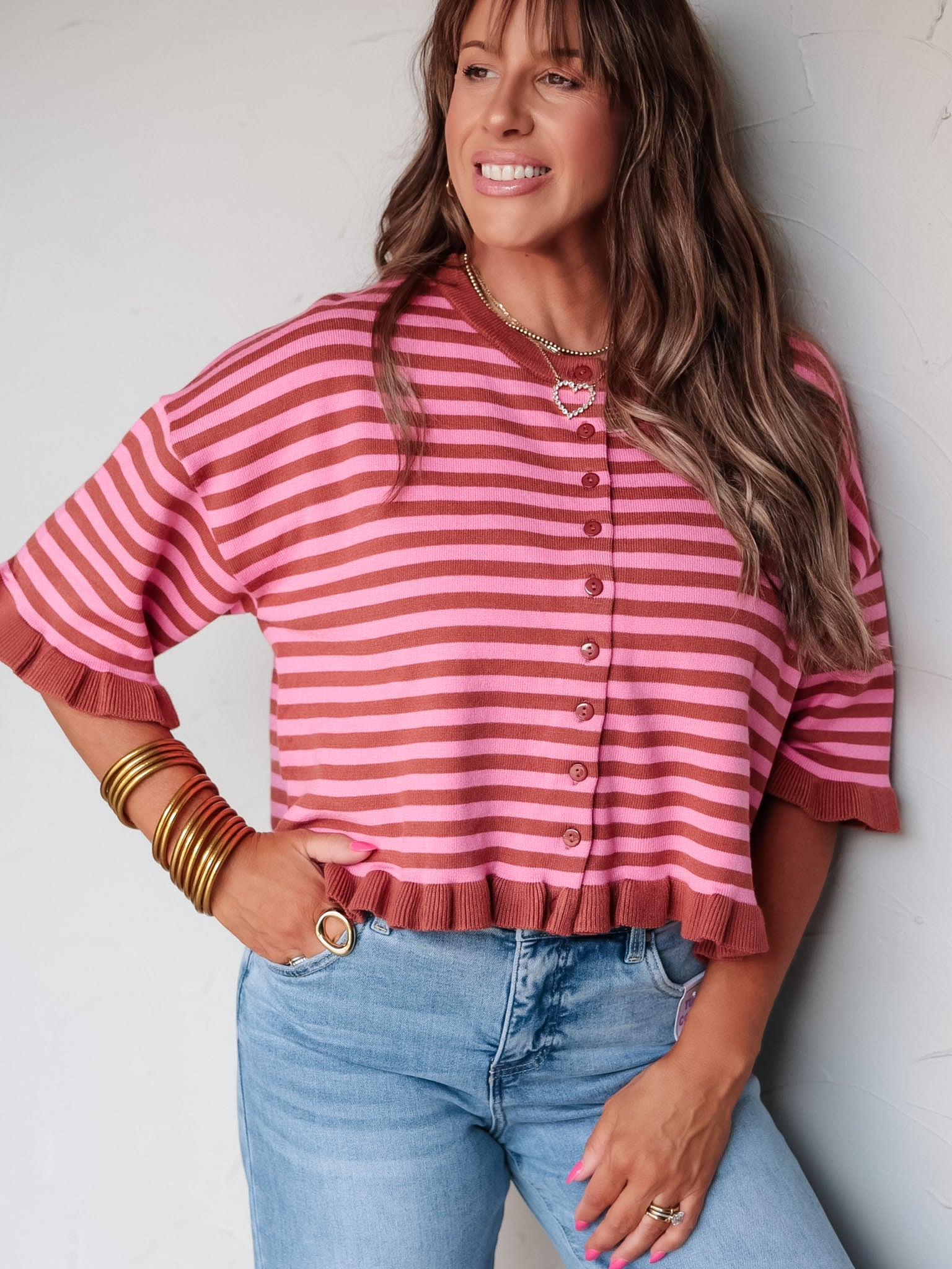 A three-quarter length photograph of a smiling woman with medium-length brown hair and bangs posing with one hand near her pocket against a textured white wall. She is wearing a short-sleeved pink and brown horizontal striped knit cardigan made from a 50% viscose, 30% polyester, and 20% nylon blend, featuring button closures down the front and ribbed ruffle trim along the cuffs and bottom hem, paired with light-wash denim jeans and styled with layered pendant necklaces and stacked gold bangles.