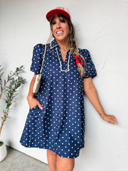 A woman smiling and leaning against a textured off-white wall with one hand in her pocket, wearing a navy blue and white polka dot shift dress crafted from a 100% cotton fabric with a comfortable inner lining. The mini-length dress features a casual pointed collar, an open V-neck placket, short puffed sleeves with tiered ruffle detailing, and convenient side pockets. Crisp white contrast piping defines the edge of the collar and frames the front neckline placket. She is styled with a cream and red trucker h