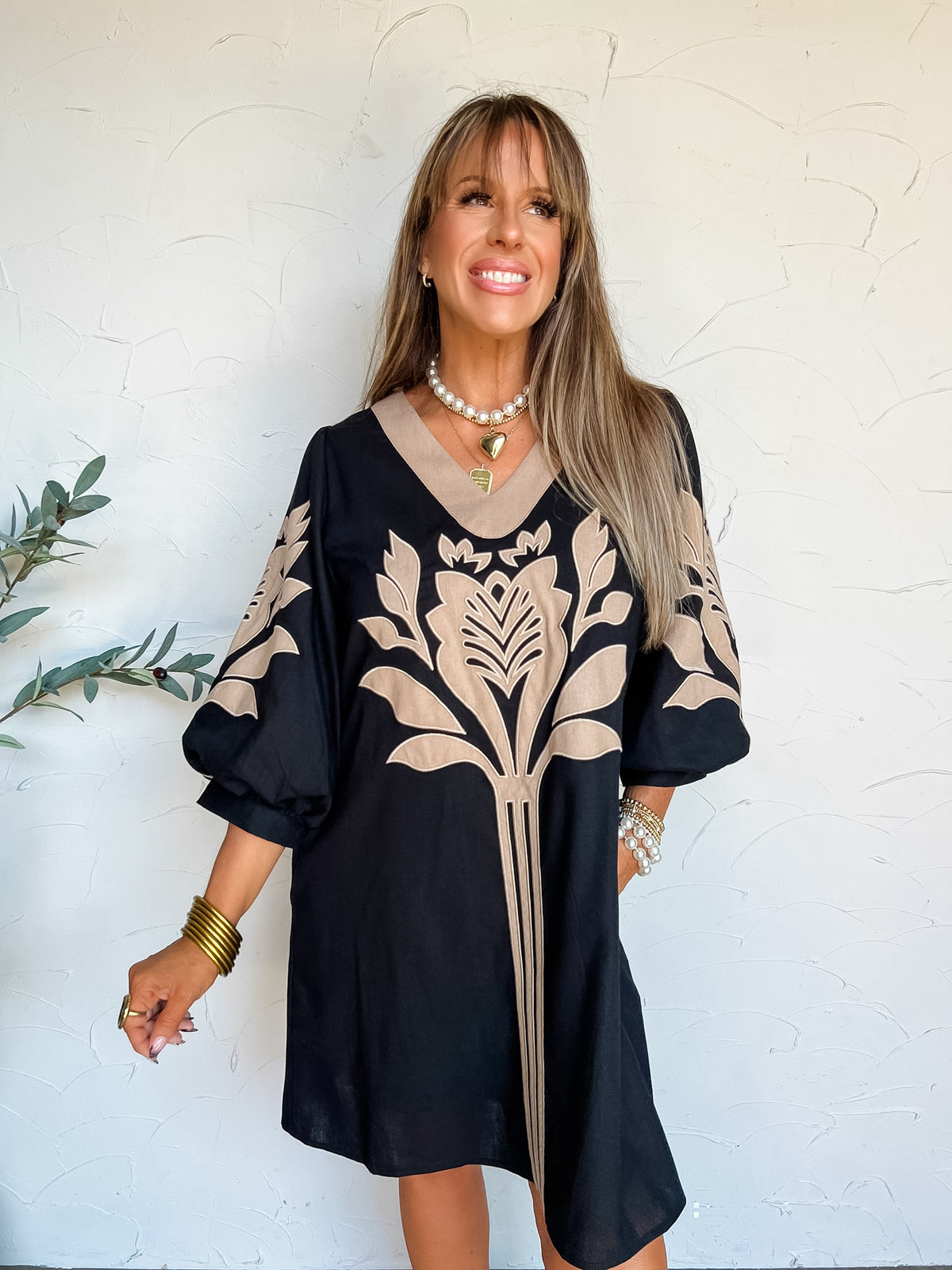 A portrait photograph of a smiling woman with straight blonde hair and bangs looking off to the side with one hand in her pocket against a textured off-white wall next to a potted olive plant. She is wearing a black knee-length shift dress made from a 70% rayon and 30% linen blend, featuring a taupe V-neck border, taupe floral leaf applique embroidery on the front bodice and sleeves, and 3/4 length balloon sleeves, styled with a white pearl choker, a gold heart pendant necklace, stacked gold bangles, and pe