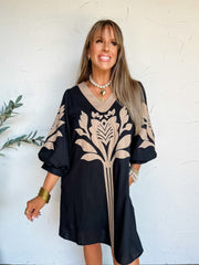 A portrait photograph of a smiling woman with straight blonde hair and bangs looking off to the side with one hand in her pocket against a textured off-white wall next to a potted olive plant. She is wearing a black knee-length shift dress made from a 70% rayon and 30% linen blend, featuring a taupe V-neck border, taupe floral leaf applique embroidery on the front bodice and sleeves, and 3/4 length balloon sleeves, styled with a white pearl choker, a gold heart pendant necklace, stacked gold bangles, and pe