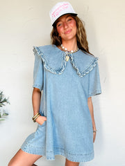 Woman wearing a denim dress with a ruffle collar and pink cap against a white wall.