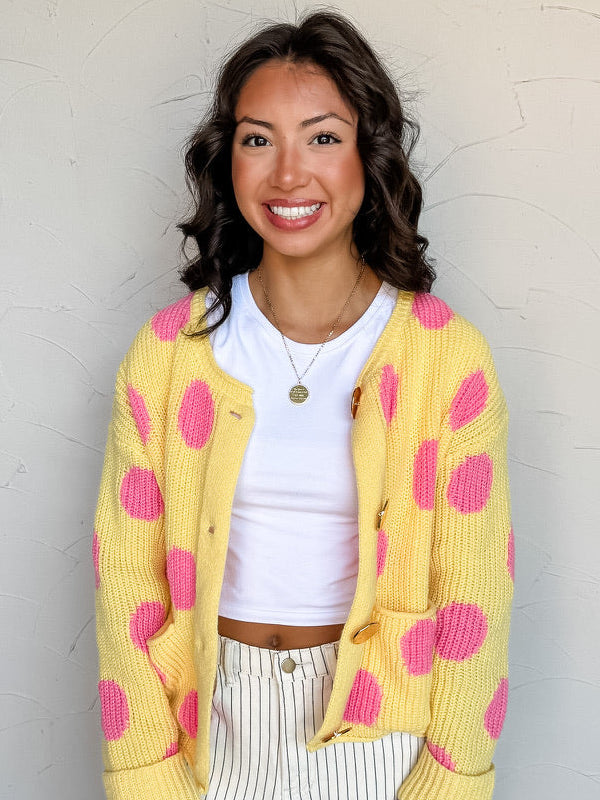 A waist-up photograph of a smiling woman with dark wavy hair looking off-camera against a textured white wall. She is wearing a yellow knit cardigan cardigan made from 100% polyester fabric, decorated with large hot pink polka dots, featuring front patch pockets, large tortoiseshell buttons, and wide ribbed cuffs, worn unbuttoned over a fitted white crop top and paired with white pinstriped jeans, styled with a round gold pendant necklace.