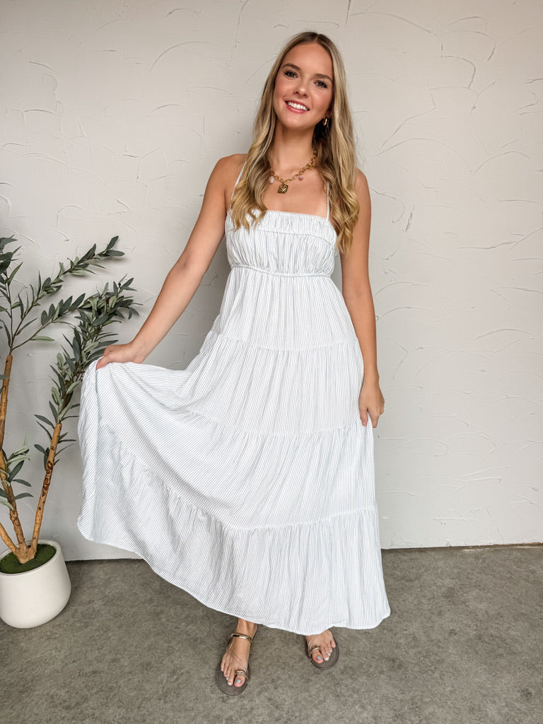 A woman smiling and posing while holding out the side of her skirt against a textured off-white wall, wearing a maxi-length dress crafted from a white and light blue 90% rayon and 10% nylon blend fabrication with a comfortable inner lining. The dress features a fine vertical blue pinstripe pattern, a straight neckline with thin adjustable cami straps, a stretchy smocked bodice, and a flowing tiered skirt. She is styled with a gold link chain necklace featuring a square padlock pendant.