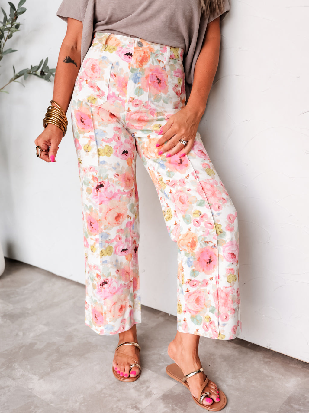 A photograph focusing from the waist down of a person leaning against a textured white wall. They are wearing high-waisted wide-leg 100 % cotton pants featuring a vibrant pink, orange, yellow, and green floral print on a cream background, designed with front patch pockets and raised center seams, styled with a tucked-in taupe short-sleeved top, gold bangles, and flat metallic strap sandals. 