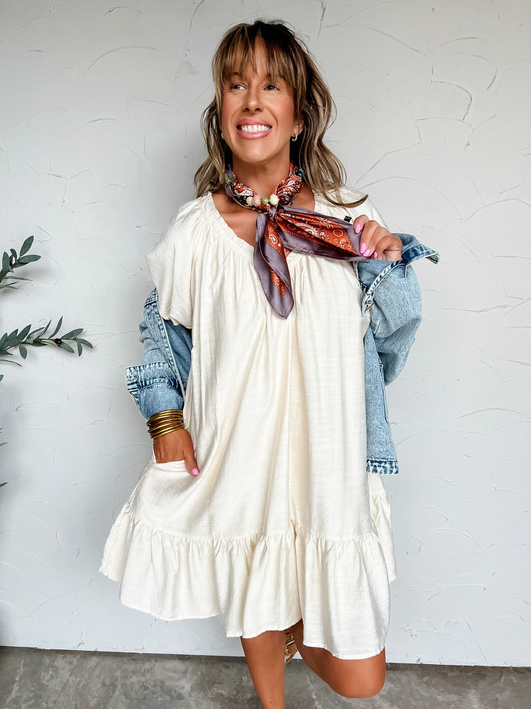 A three-quarter length photograph of a smiling woman with long brown hair and bangs posing against a textured off-white wall while holding a patterned neck scarf. She is wearing an oversized short-sleeved cream babydoll mini dress made from an 80% rayon and 20% nylon blend, featuring a gathered round neckline, side pockets, and a ruffled hem, layered with a light-wash denim jacket worn off the shoulders, styled with a grey and rust orange paisley neck scarf tied with beaded accent slides and stacked gold ba
