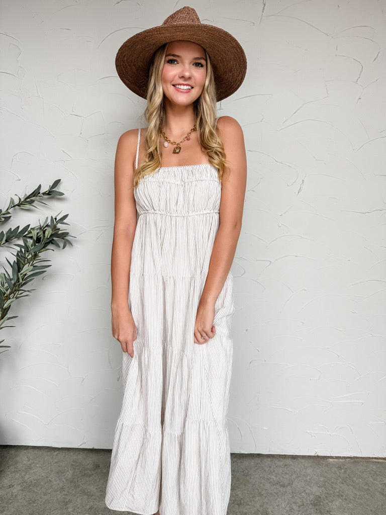 A woman smiling and posing while holding out the side of her skirt against a textured off-white wall, wearing a maxi-length dress crafted from a mocha-colored 90% rayon and 10% nylon blend fabrication with a comfortable inner lining. The mocha dress features a fine vertical pinstripe pattern, a straight neckline with thin adjustable cami straps, a stretchy smocked bodice, and a flowing tiered skirt. She is styled with a wide-brimmed brown straw hat.