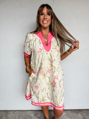 A front-facing photograph of a smiling woman with straight light-brown hair and front bangs holding a strand of her hair and placing one hand in her dress pocket against a textured white wall. She is wearing a relaxed-fit mini dress made from 100% cotton, featuring a soft cream background printed with pink blossoms and olive green botanical stems, a bright hot pink ribbed V-neckline with white trim, short sleeves with hot pink cuffs, a matching pink border along the hem, and side pockets, styled with stacke