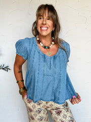 A three-quarter length photograph of a smiling woman with long brown hair and bangs posing against a textured off-white wall. She is wearing a short-sleeved medium-wash blue denim blouse made from 100% Tencel, featuring a wide scoop neckline with a small central split slit, short elasticized puff sleeves, and decorative vertical seam panel stitching, paired with cream-colored pants printed with a maroon and floral medallion pattern, styled with a colorful beaded necklace, gold pendant, and stacked brass ban