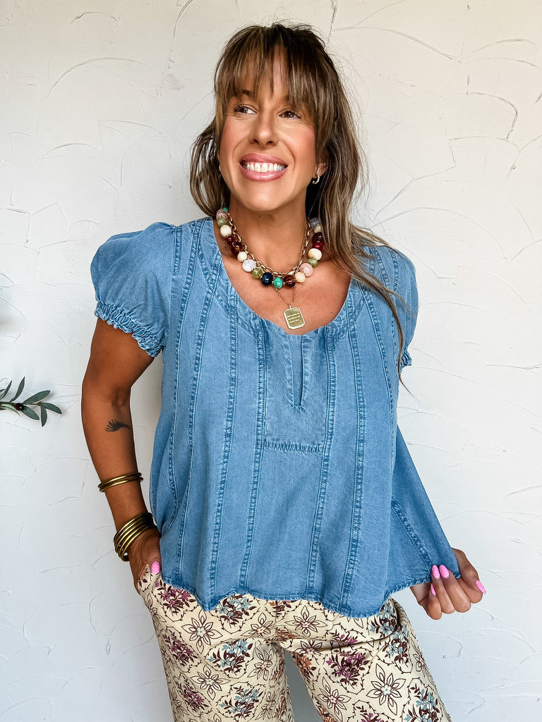 A three-quarter length photograph of a smiling woman with long brown hair and bangs posing against a textured off-white wall. She is wearing a short-sleeved medium-wash blue denim blouse made from 100% Tencel, featuring a wide scoop neckline with a small central split slit, short elasticized puff sleeves, and decorative vertical seam panel stitching, paired with cream-colored pants printed with a maroon and floral medallion pattern, styled with a colorful beaded necklace, gold pendant, and stacked brass ban