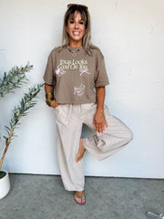 Woman wearing a brown t-shirt with text and beige pants against a white wall.