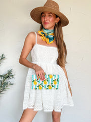 Woman in a white dress with a straw hat and colorful bag, standing against a white wall.