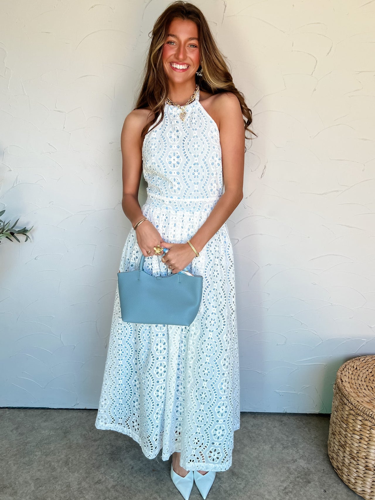 Woman in a white eyelet dress holding a blue handbag against a white wall.