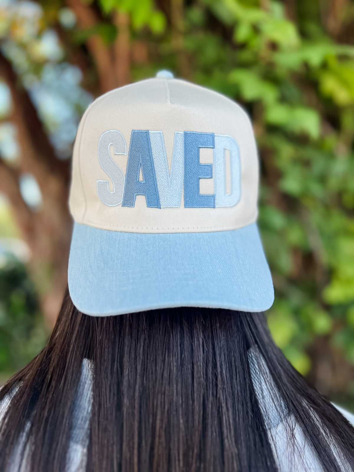 A person viewed from behind wearing a baseball cap with a cream crown and light blue brim, featuring the word 'SAVED' embroidered in light blue and gray on the front. The background shows a blurred outdoor scene with green foliage.