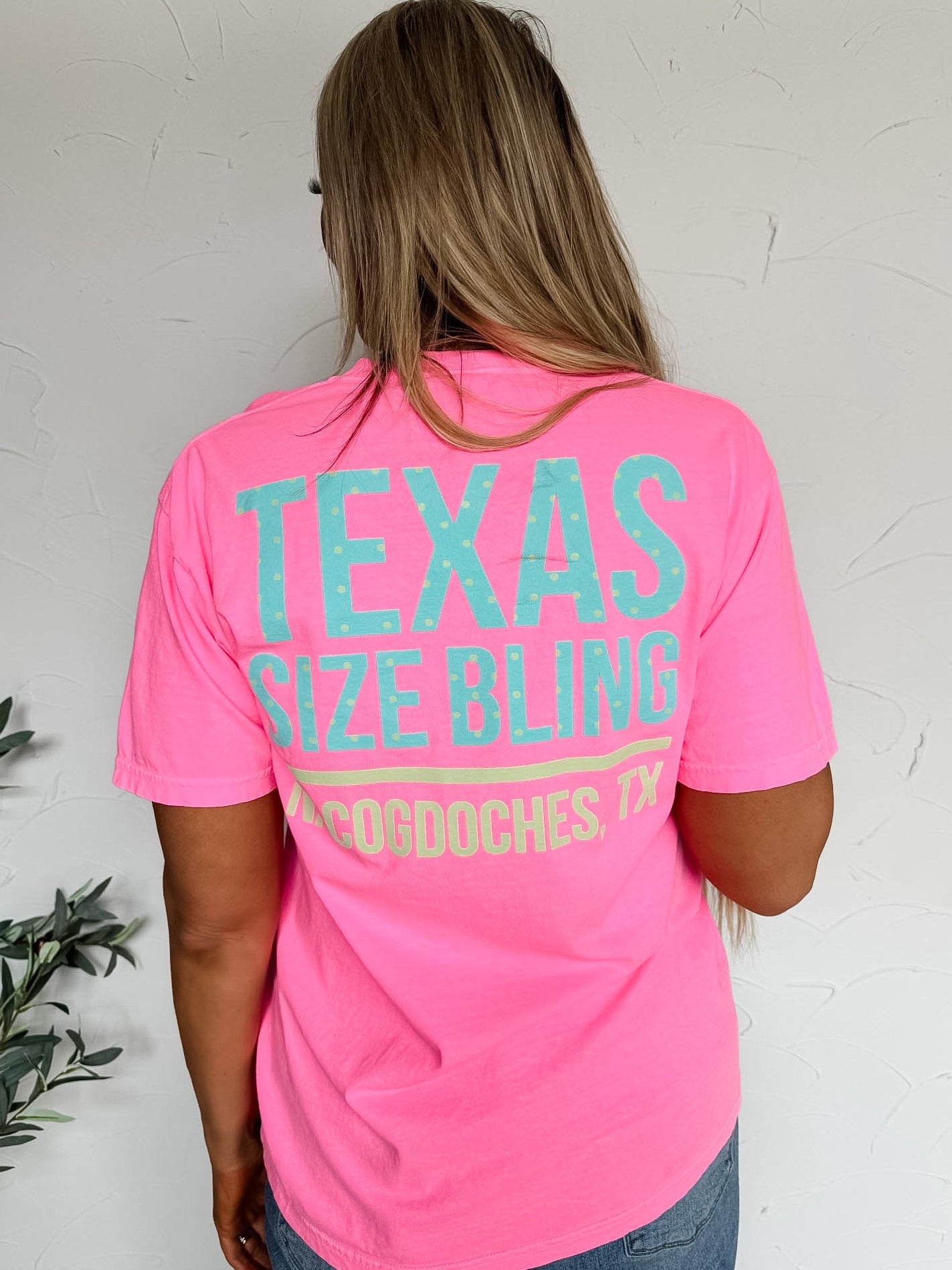 Person wearing a pink t-shirt with 'Texas Size Bling' text on the back against a white wall.