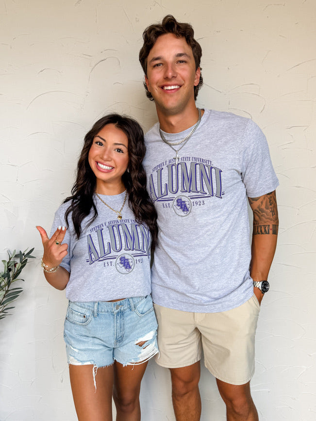 Two people wearing 'Alumni' shirts standing indoors with a white wall background.