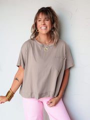 A waist-up photograph of a smiling woman with medium-length brown hair and bangs posing with her arms spread wide against a textured white wall. She is wearing a taupe brown short-sleeved oversized t-shirt made from 100% cotton fabric featuring a chest pocket and drop shoulders, paired with bright pink pants and styled with gold hoop earrings, layered gold pendant necklaces, and stacked gold bangles.