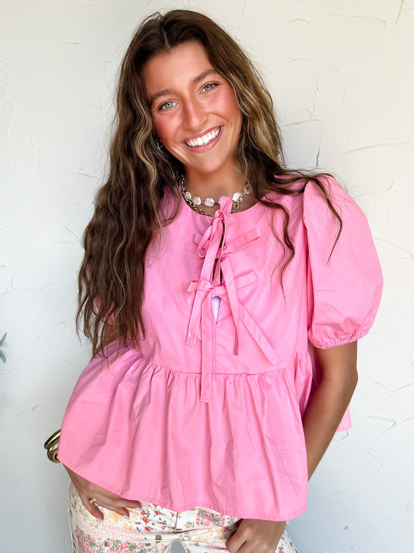 Woman wearing a pink peplum top with bow details with puff sleeves against a white wall.
