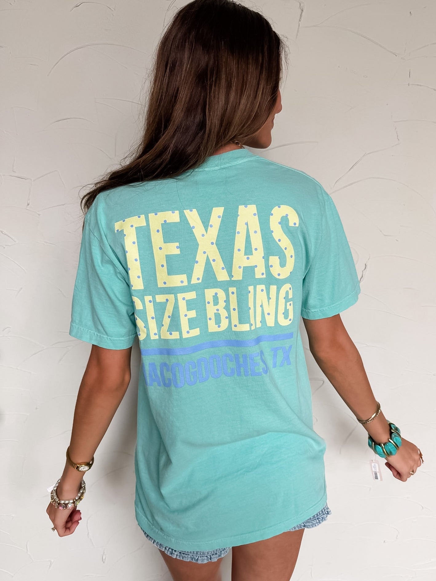 Person wearing a teal t-shirt with 'TEXAS SIZE BLING' text on the back against a white wall.