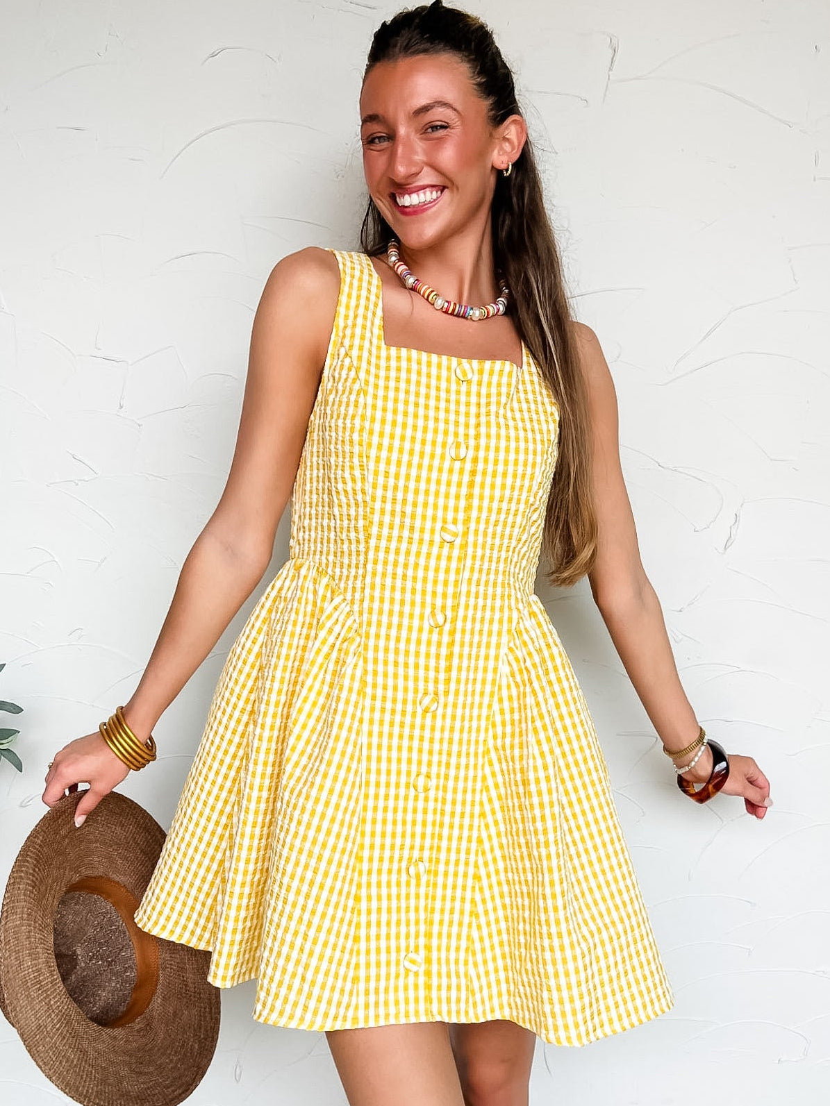 Woman wearing a yellow checkered dress holding a straw hat against a white wall.