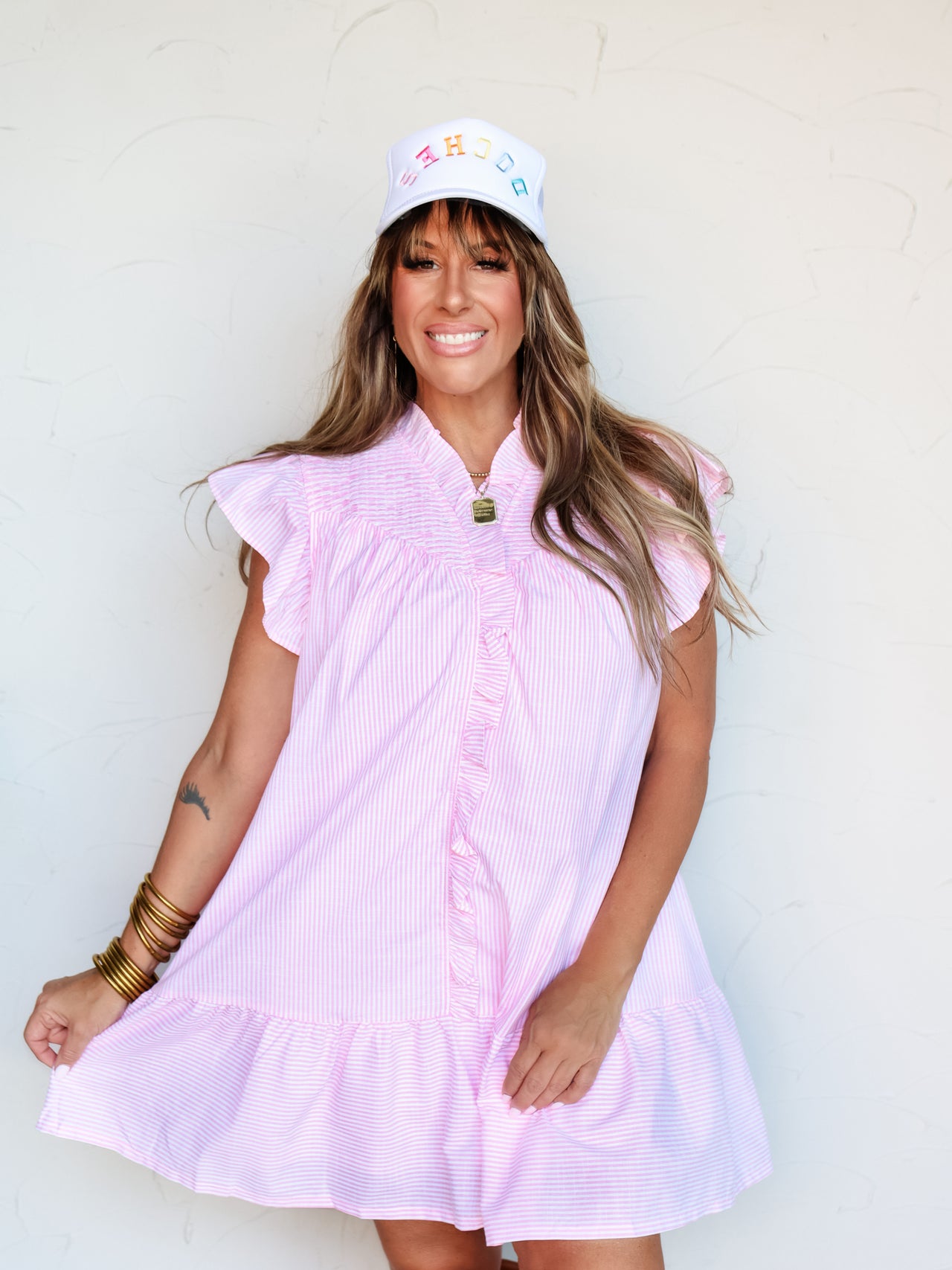 A full-length photograph of a smiling brunette woman with bangs posing dynamically against a textured white wall while holding the edge of her skirt. She is wearing a relaxed pink and white vertically striped short-sleeve dress made from a 75% cotton and 25% polyester blend fabric, featuring a ruffled collar, ruffle-trimmed front placket, flutter sleeves, and a tiered hemline, styled with a white trucker hat and gold bangles stacked on her wrist.