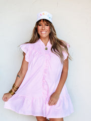 A full-length photograph of a smiling brunette woman with bangs posing dynamically against a textured white wall while holding the edge of her skirt. She is wearing a relaxed pink and white vertically striped short-sleeve dress made from a 75% cotton and 25% polyester blend fabric, featuring a ruffled collar, ruffle-trimmed front placket, flutter sleeves, and a tiered hemline, styled with a white trucker hat and gold bangles stacked on her wrist.