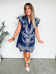Woman wearing a blue dress with white patterns against a white wall.
