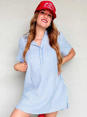 Woman wearing a blue and white striped dress with a red cap and shoes against a white wall.