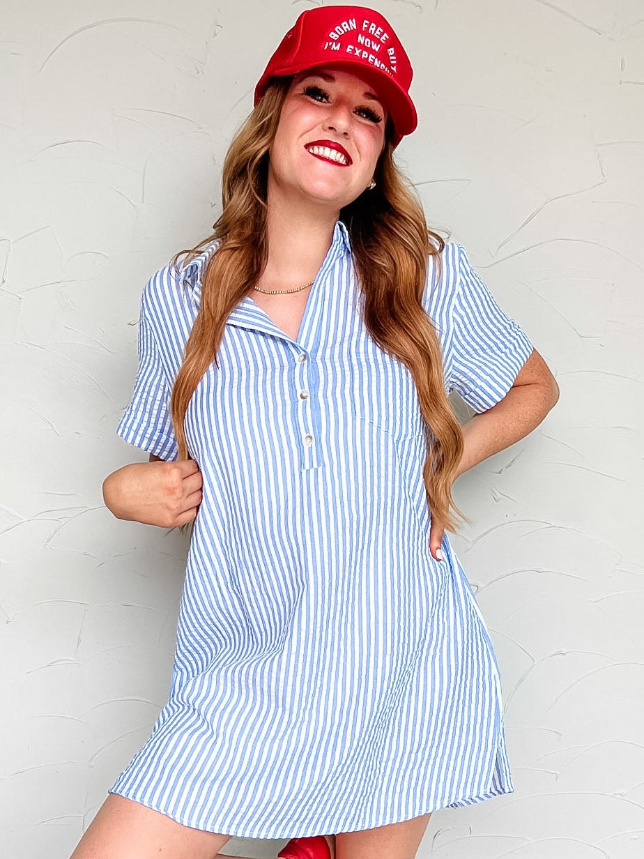 Woman wearing a blue and white striped dress with a red cap and shoes against a white wall.