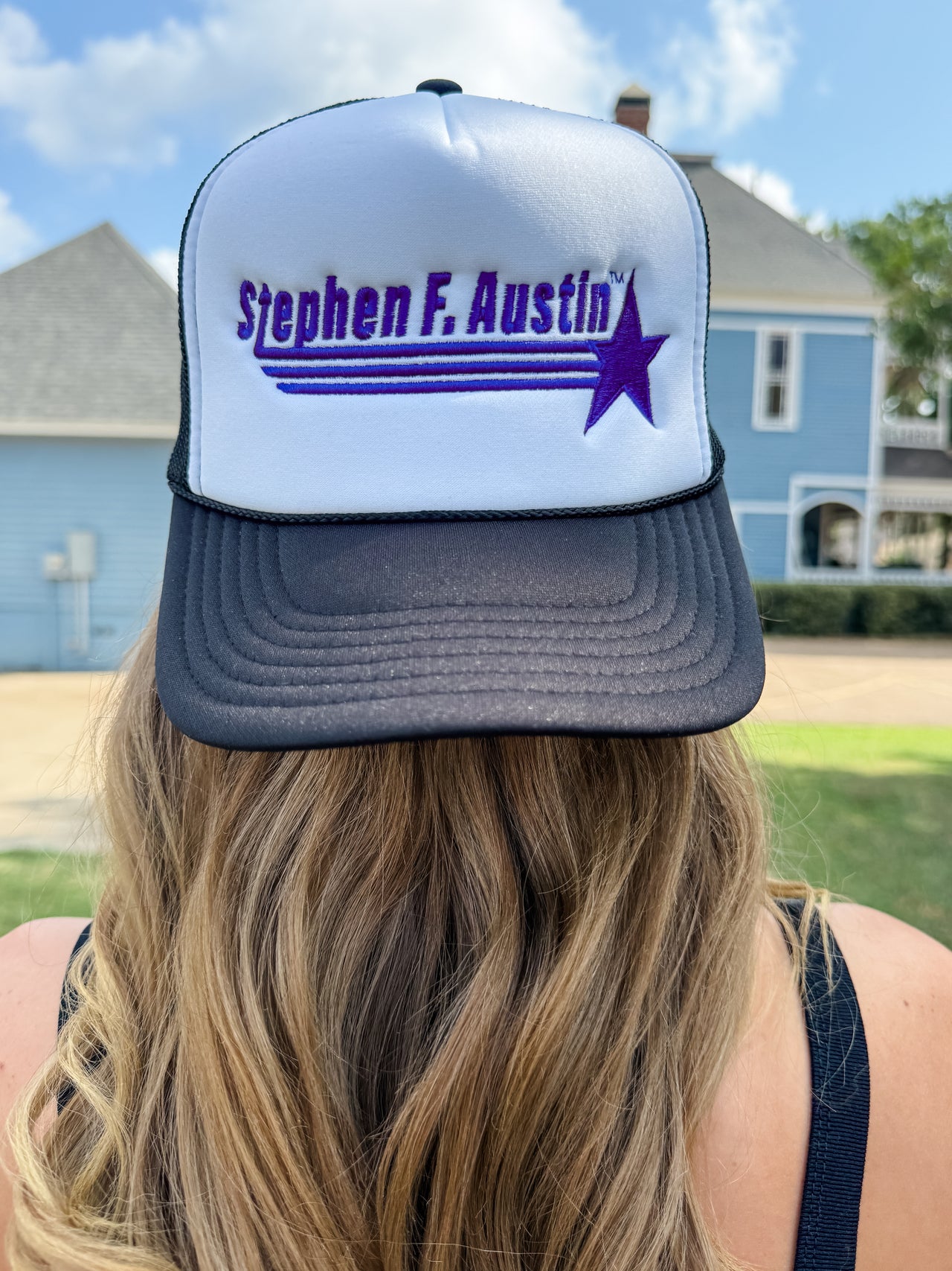 A person viewed from behind wearing a white and dark gray trucker hat with purple embroidered text reading 'Stephen F. Austin' and a purple star graphic, outdoors in front of houses and a blue sky.