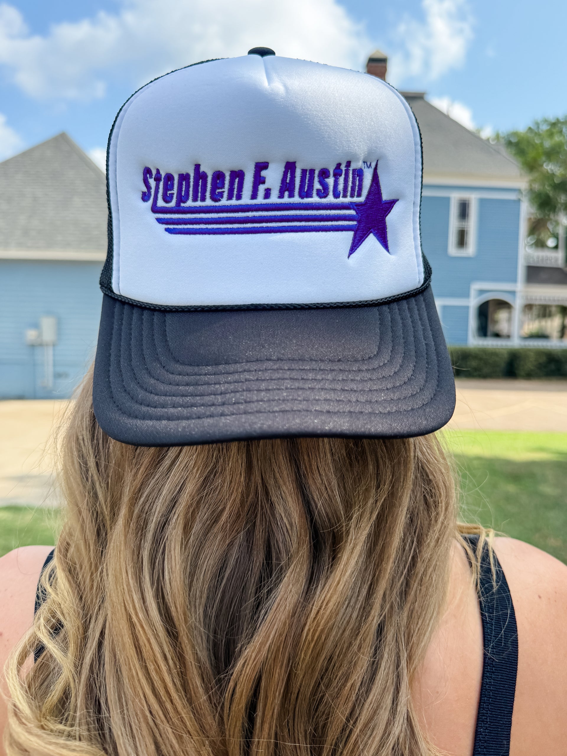 A person viewed from behind wearing a white and dark gray trucker hat with purple embroidered text reading 'Stephen F. Austin' and a purple star graphic, outdoors in front of houses and a blue sky.