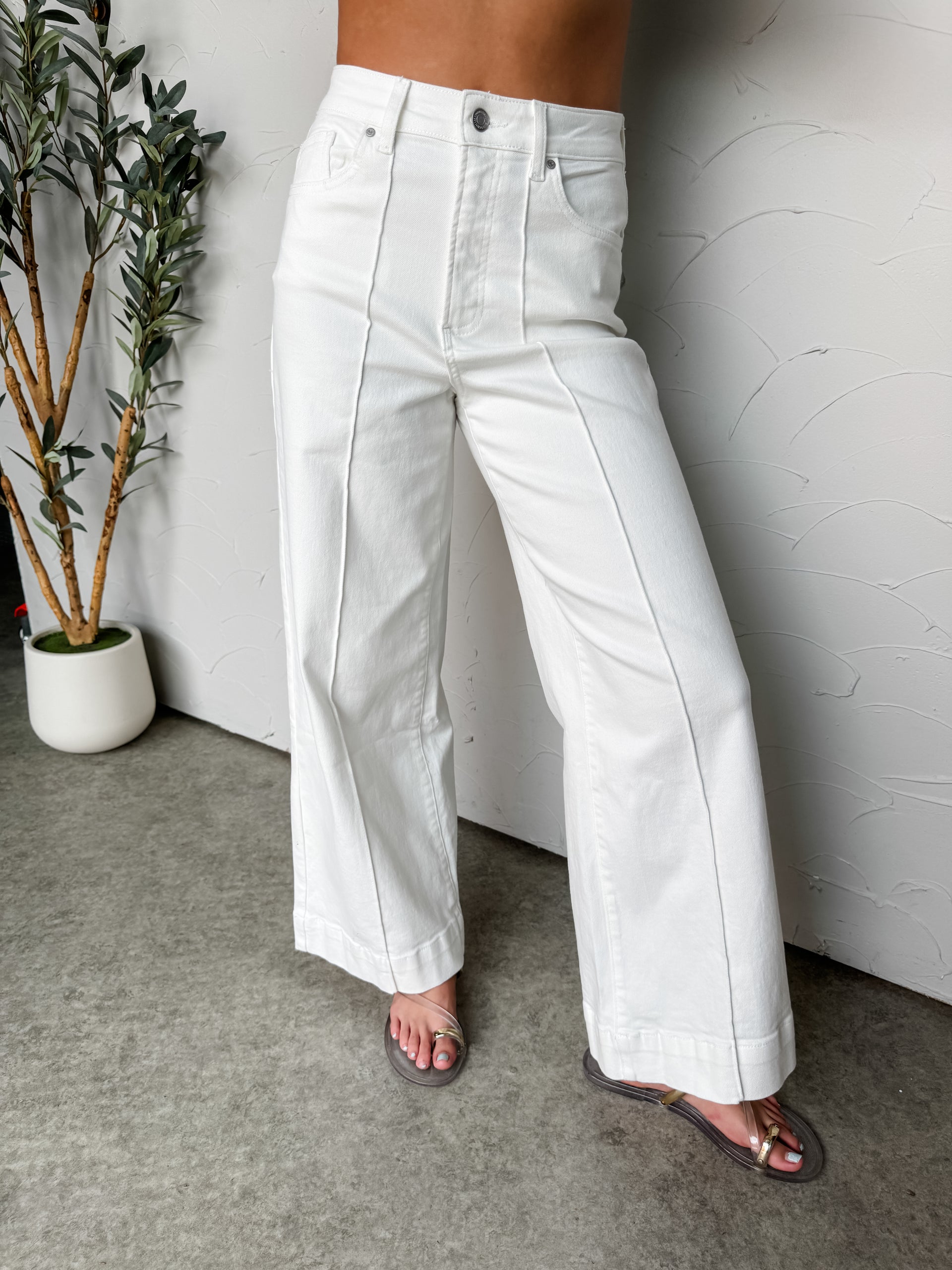 Person wearing white wide-leg pants standing against a white wall with a plant in the corner.