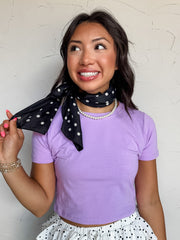 A three-quarter length photograph of a smiling woman with dark wavy hair holding a strand of her hair while posing against a textured white wall. She is wearing a fitted lavender purple short-sleeved cropped t-shirt made from a 95% cotton and 5% spandex blend, paired with a white and black polka dot tiered ruffled mini skirt and styled with a black polka dot neck scarf, pearl necklace, gold beaded bracelets, and black cowboy boots.