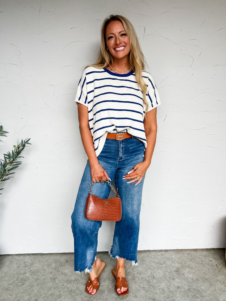 Woman wearing a striped shirt, blue jeans, and holding a brown handbag against a white wall.