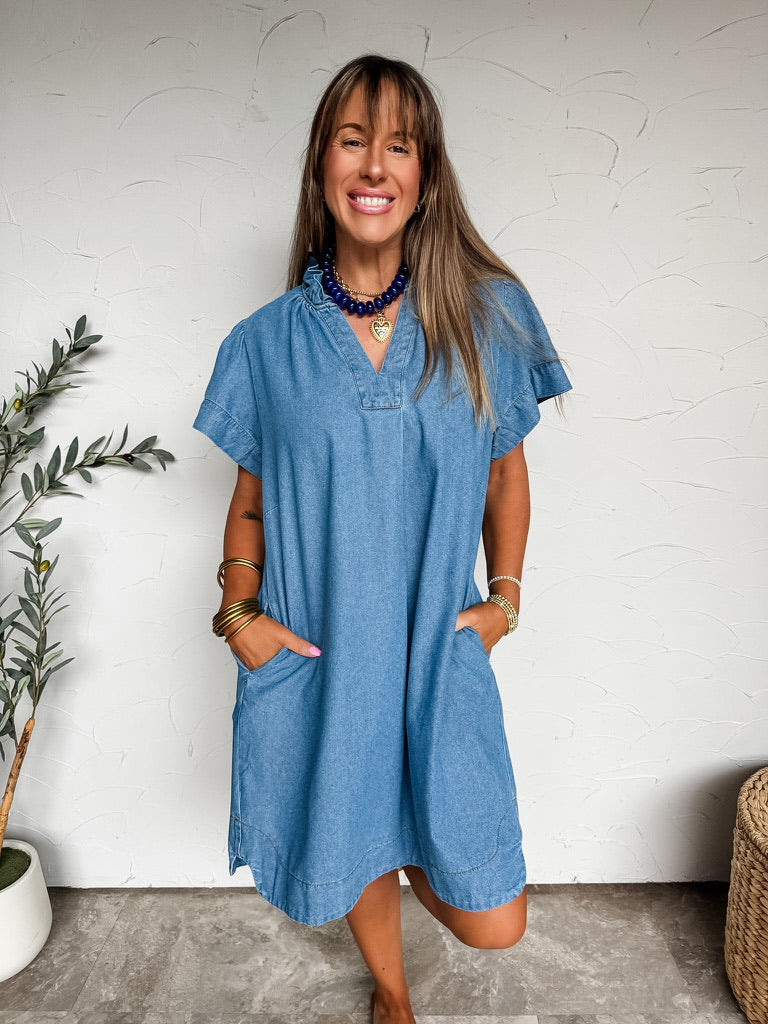 A full-length photograph of a smiling woman with straight brown hair and bangs posing with both hands in her front pockets against a textured white wall. She is wearing a short-sleeved medium wash blue denim mini shift dress made from an 85% cotton, 10% polyester, and 5% viscose blend, featuring a ruffled high collar with a split V-neckline, short flutter sleeves, large curved front patch pockets, and a relaxed knee-length hem, styled with a chunky navy blue beaded necklace, a gold heart pendant, stacked go