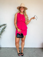 A smiling blonde woman stands against an off-white wall wearing a vibrant magenta pink two-piece matching set made from 100% polyester. The set includes a sleeveless tailored vest with a round neckline, a button-down front featuring gold-rimmed buttons, and decorative welt pockets, paired with coordinating high-waisted tailored shorts. She is styled with a wide-brimmed straw hat, stacked gold bangles, a small black shoulder bag with a gold chain strap, and black flat sandals with a large gold medallion.
