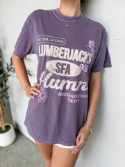 Person wearing a purple t-shirt with 'Lumberjacks' text and graphics, standing against a white wall.