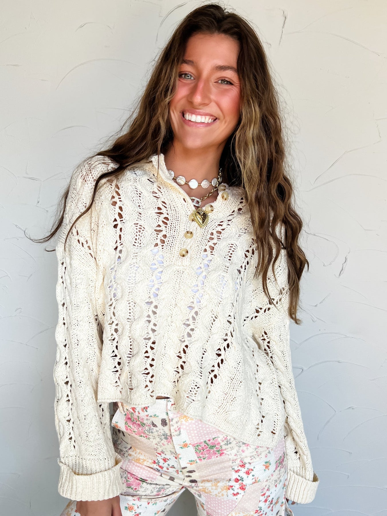Woman wearing a cream lace eyelet sweater over a white top with floral pants against a white background