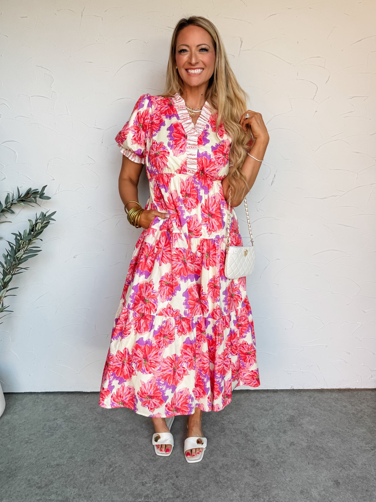 A smiling blonde woman stands against an off-white wall, wearing a tiered floral maxi dress. The dress is made from a 100% cotton fabric with lining and features an all-over bold floral print in shades of coral pink, purple, and cream, detailed with a white ruffle-trimmed V-neckline and short puff sleeves. She has her hand in her pocket and is accessorized with stacked gold bangles, layered necklaces, a small white quilted crossbody bag, and white flat sandals.