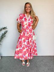 A smiling blonde woman stands against an off-white wall, wearing a tiered floral maxi dress. The dress is made from a 100% cotton fabric with lining and features an all-over bold floral print in shades of coral pink, purple, and cream, detailed with a white ruffle-trimmed V-neckline and short puff sleeves. She has her hand in her pocket and is accessorized with stacked gold bangles, layered necklaces, a small white quilted crossbody bag, and white flat sandals.