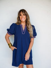 A full-length photograph of a smiling brunette woman with bangs posing with one hand in her pocket against a textured white wall. She is wearing a relaxed navy blue shift dress made from a 60% viscose and 40% polyester blend fabric, featuring a split V-neckline and puff short sleeves detailed with hot pink geometric embroidery, styled with gold bangles stacked on her wrist.