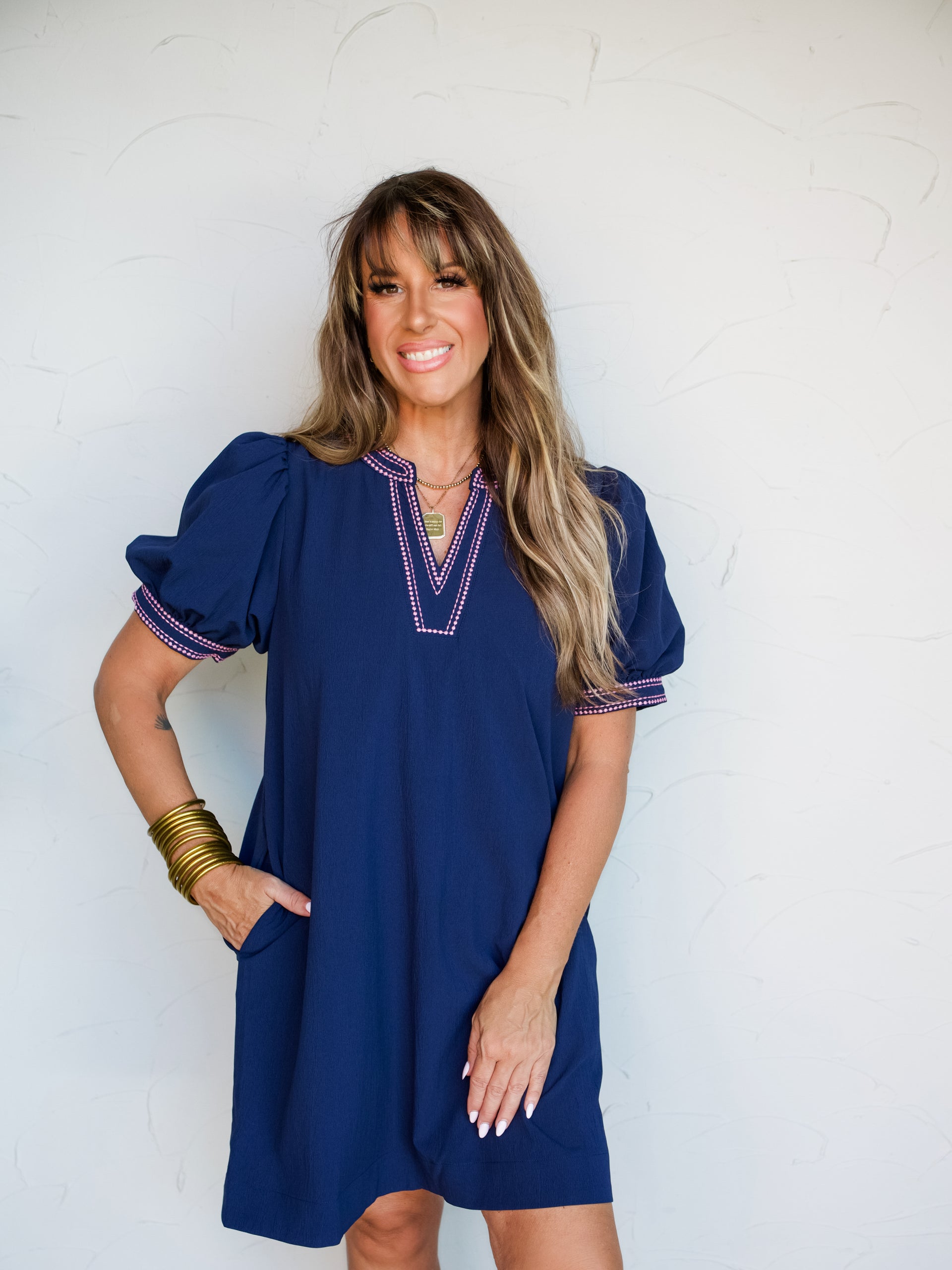 A full-length photograph of a smiling brunette woman with bangs posing with one hand in her pocket against a textured white wall. She is wearing a relaxed navy blue shift dress made from a 60% viscose and 40% polyester blend fabric, featuring a split V-neckline and puff short sleeves detailed with hot pink geometric embroidery, styled with gold bangles stacked on her wrist.