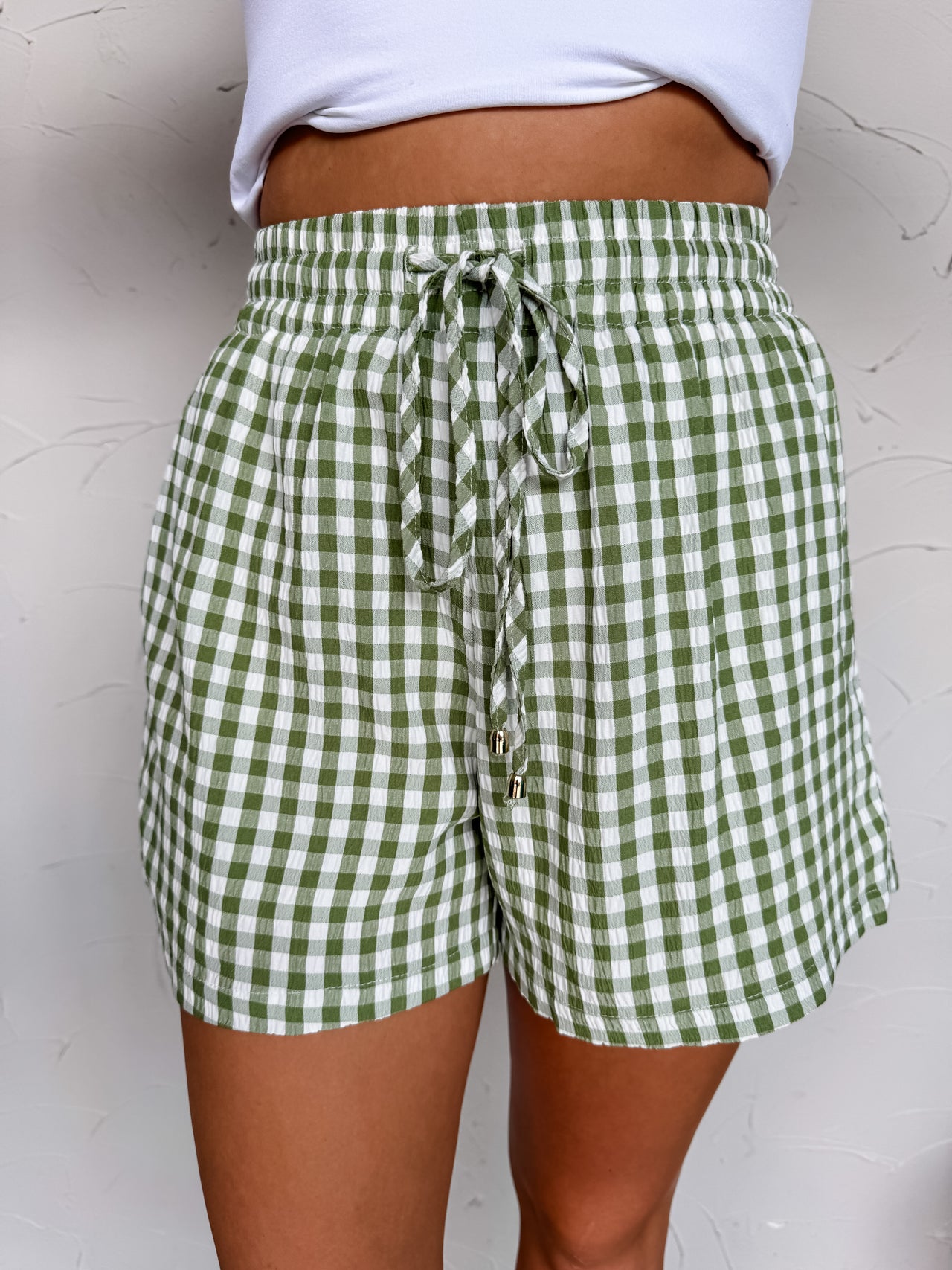 A close-up shot of a person standing against a textured off-white wall, wearing relaxed green and white gingham patterned shorts crafted from a 65% polyester and 35% cotton blend. The shorts feature a high-rise smocked elastic waistband with a matching gingham drawstring tie finished with shiny gold hardware tips. The lightweight shorts are paired with a simple white cropped top, showing a portion of the person's midriff.