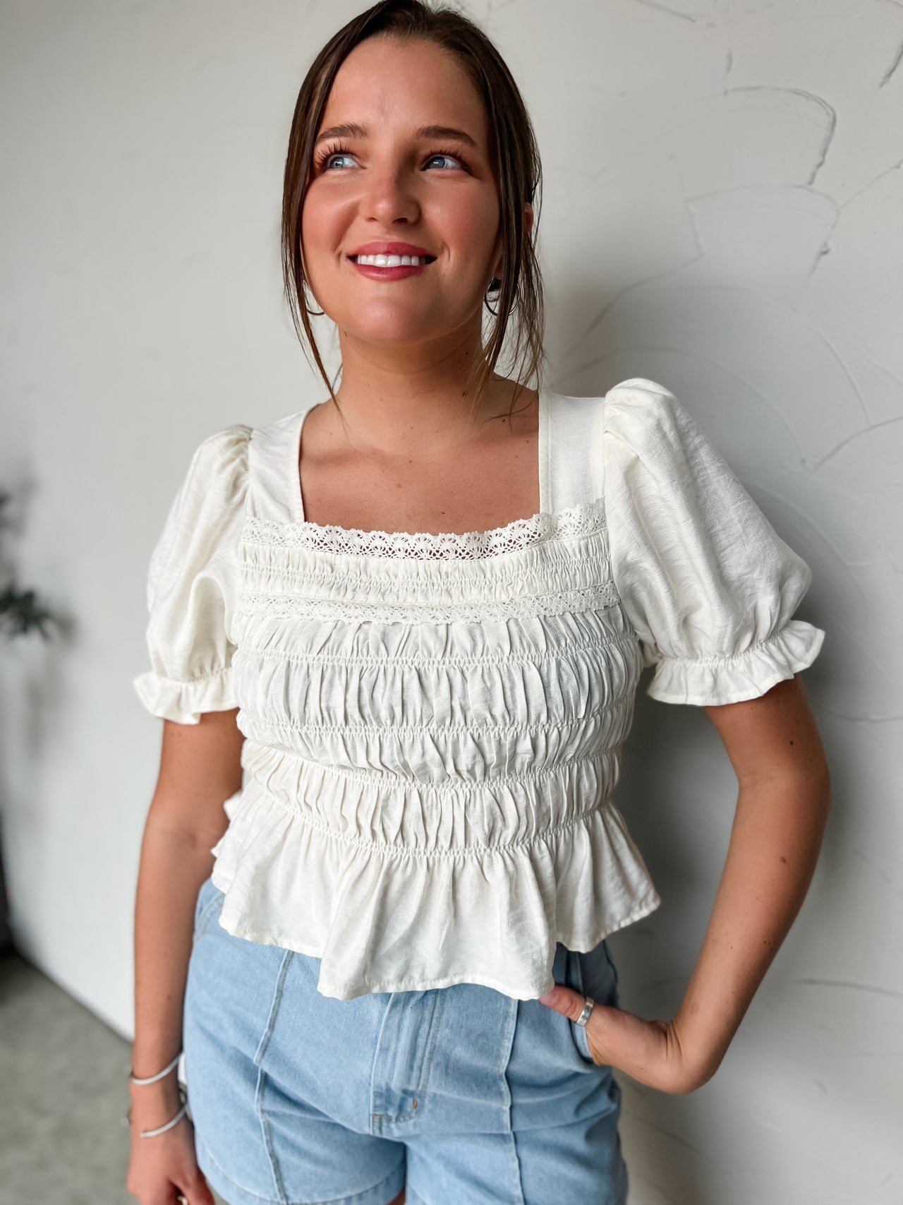 Woman wearing a white blouse with puffed sleeves and blue jeans against a light gray wall.