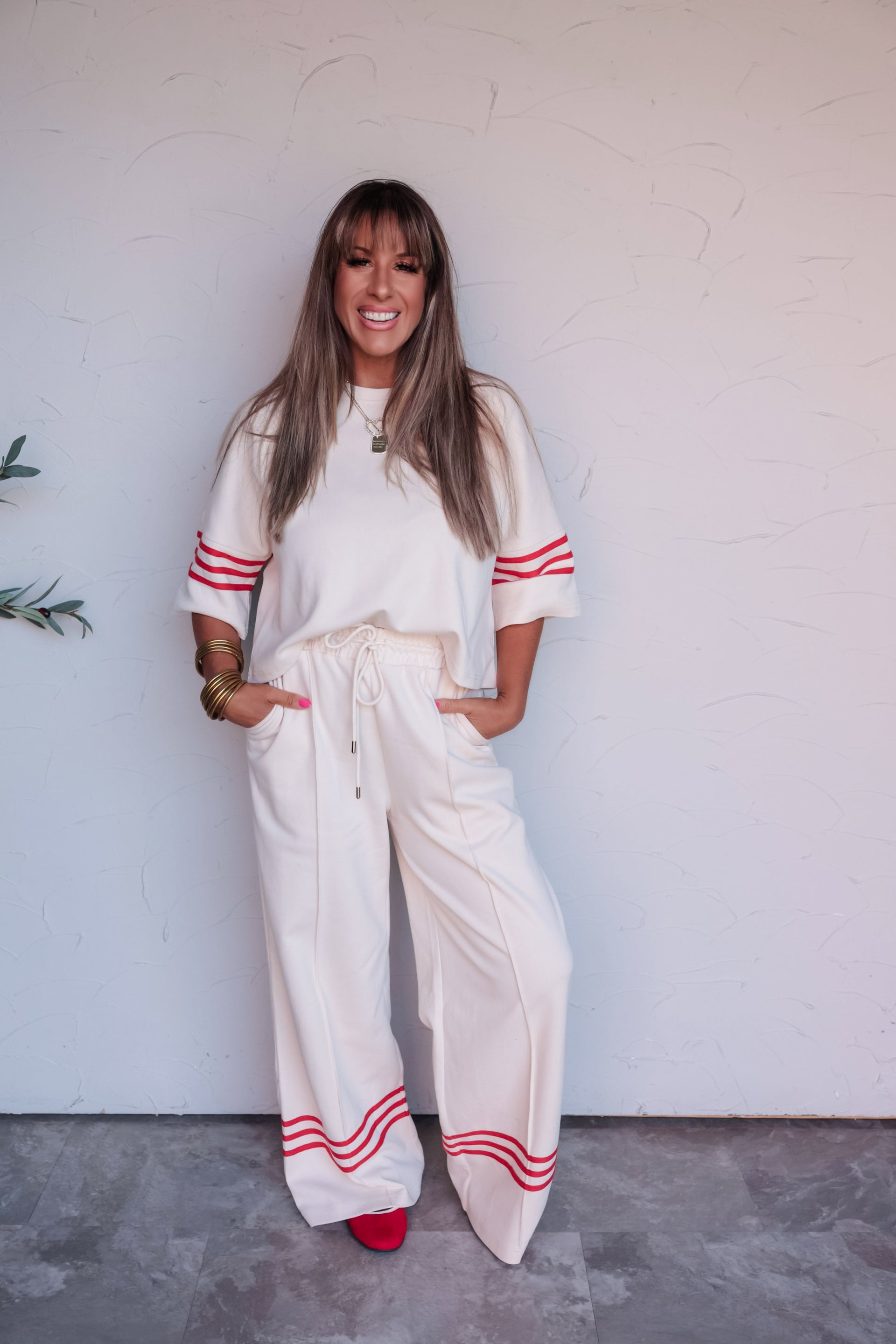 A full-length photograph of a smiling woman with straight blonde hair and bangs posing with her hands in her pockets against a textured white wall. She is wearing a cream-colored matching two-piece loungewear set featuring red athletic stripes on the short sleeves and wide-leg pant cuffs, made from a 60% cotton and 40% polyester blend fabric, styled with red shoes, gold bangles, and a gold pendant necklace.