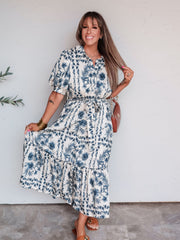 A full-length photograph of a smiling woman with long brown hair and straight bangs posing against a textured white wall. She is wearing a cream and blue floral printed maxi shirt dress made from a 70% rayon and 30% nylon blend fabric, featuring a collar, button-down bodice, puffed short sleeves, a tied waist belt, side pockets, and a tiered ankle-length skirt, styled with brown slide sandals, a cognac shoulder purse, gold bangles, and a gold necklace.