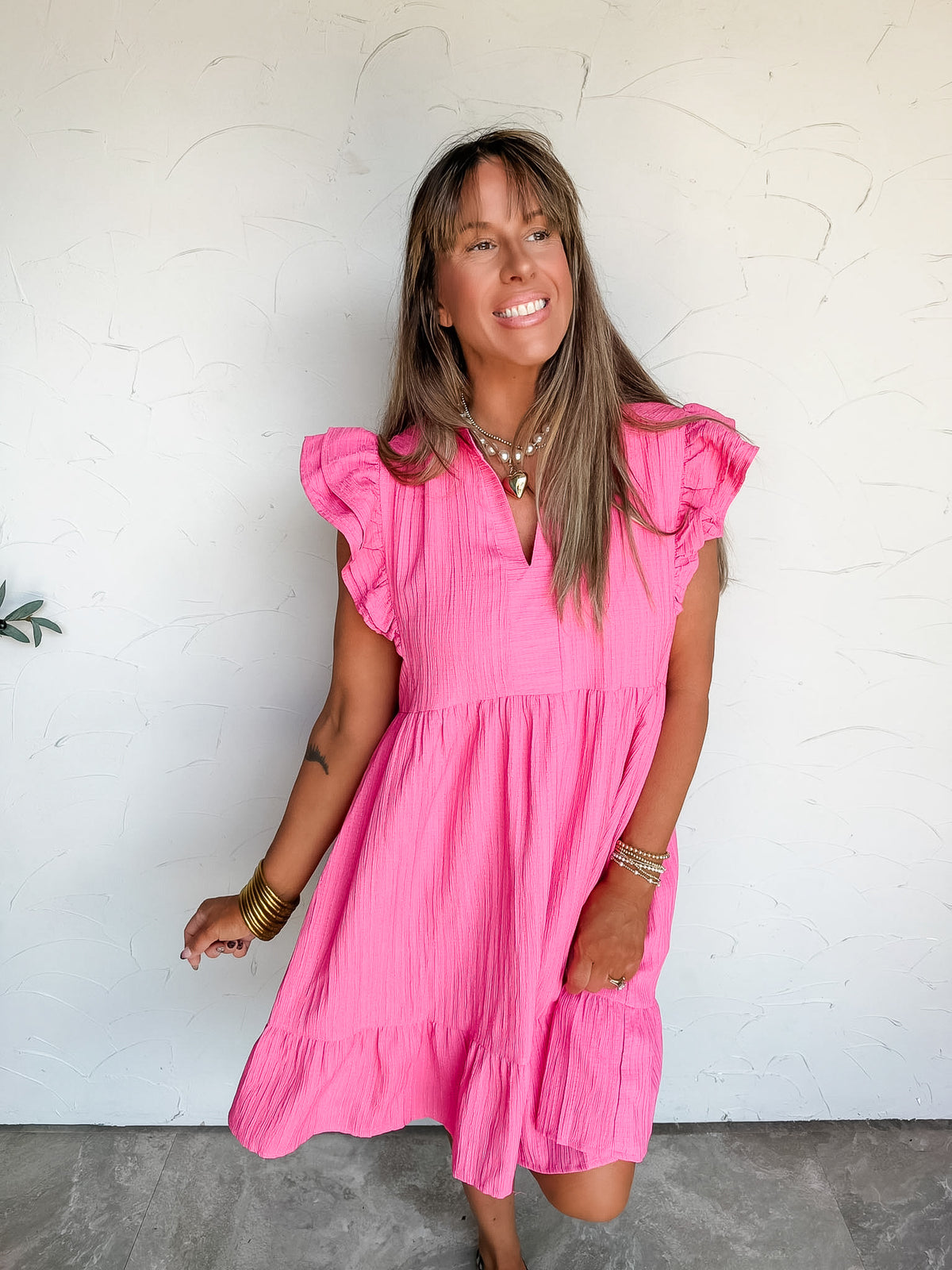 Lesly modeling the Umgee Never Waste A Moment Double Sleeve Dress in Peony, size Large, made from 100% polyester with textured pink fabric, double-layer ruffle sleeves, a split neckline with stand collar, and a gathered tiered skirt with a ruffled hem, styled with a layered pearl and heart necklace, gold-tone bracelets, and patterned ankle boots.