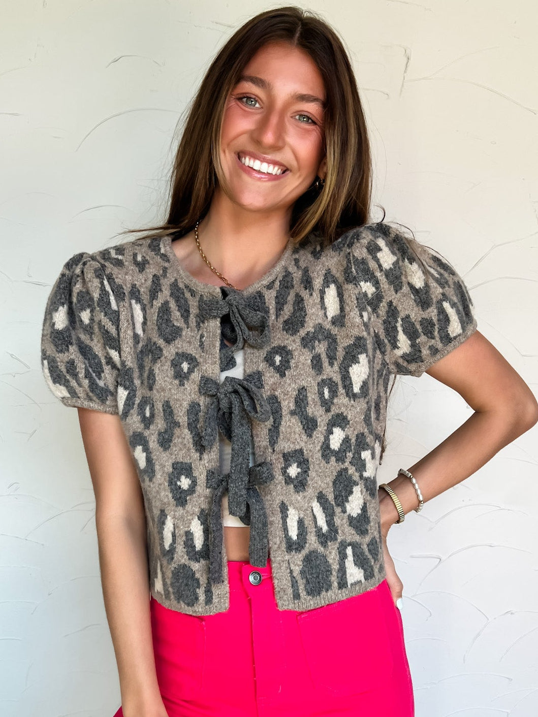 Woman wearing a leopard print cardigan bow tie top and bright pink pants against a white wall.